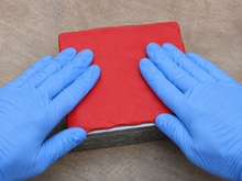 Load image into Gallery viewer, Two gloved hands pressing a 7-inch by 7-inch red intumescent fire putty pad onto a galvanized steel single-gang electrical outlet box on a workbench during installation
