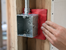 Load image into Gallery viewer, Bright red Everkem intumescent fire putty pad pressed onto the side of a galvanized steel single-gang electrical outlet box in a wood-stud wall cavity before drywall installation
